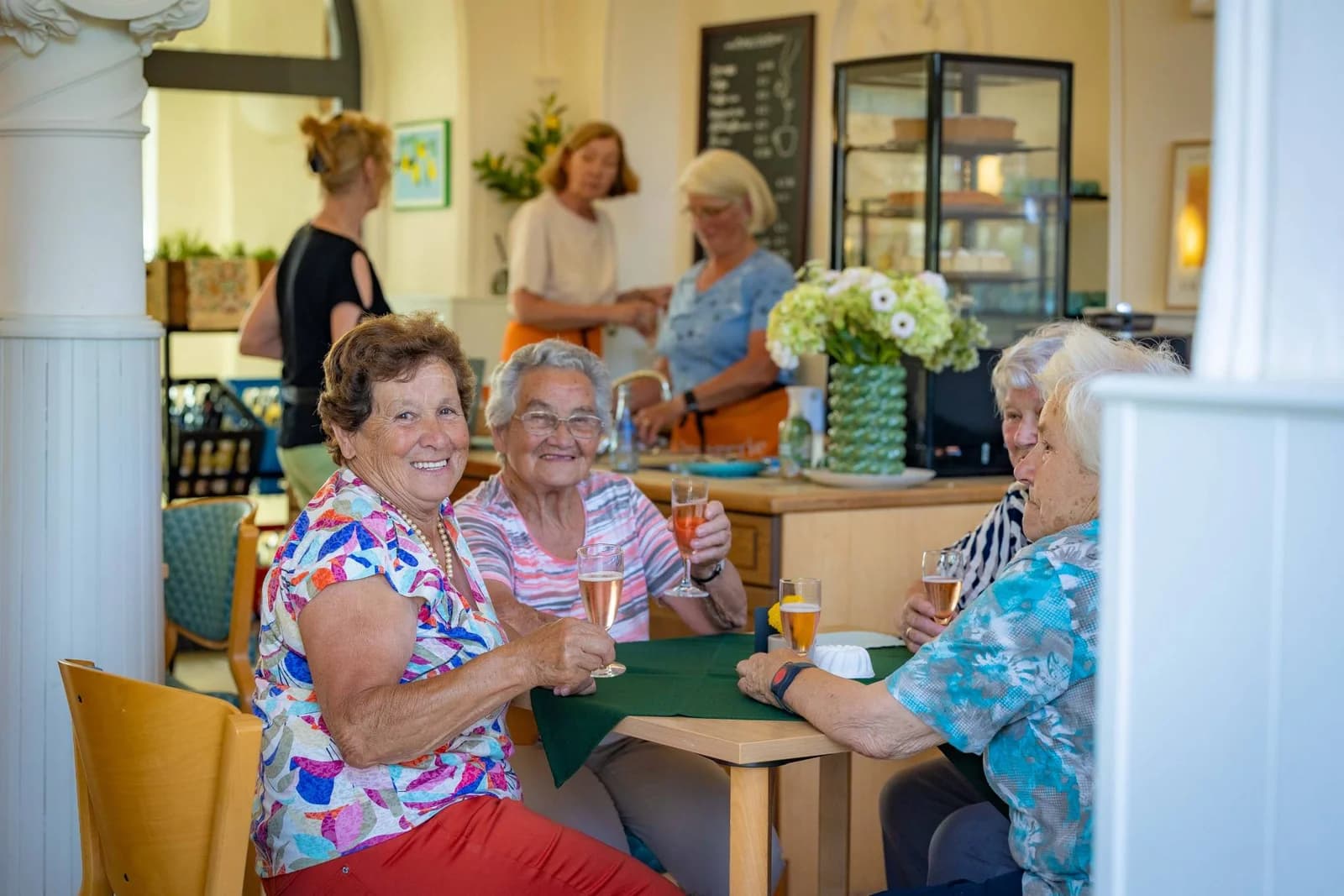 Ältere Frauen stoßen in einem Café mit ihren Getränken an, im Hintergrund arbeiten drei Frauen an der Theke