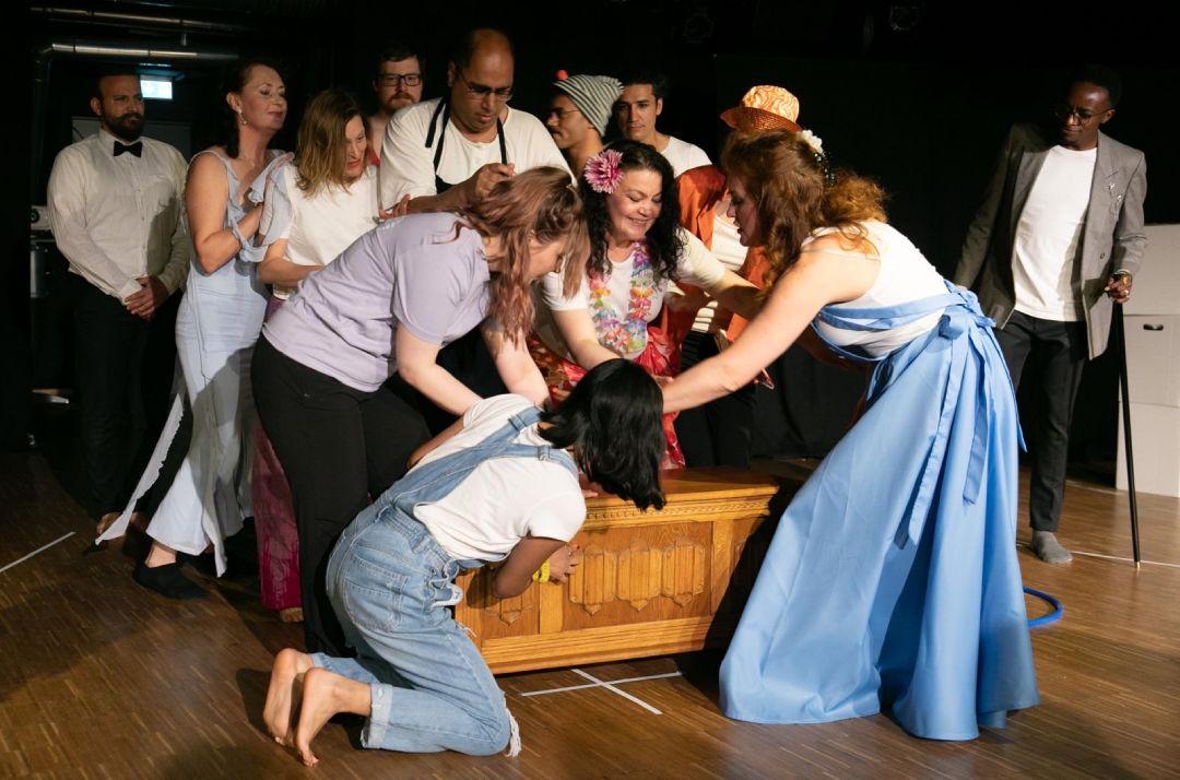 Die Theatergruppe von Campus Asyl während einer Vorführung. Eine große Gruppe ist zu sehen, die über eine Holzkommode gebeugt ist.