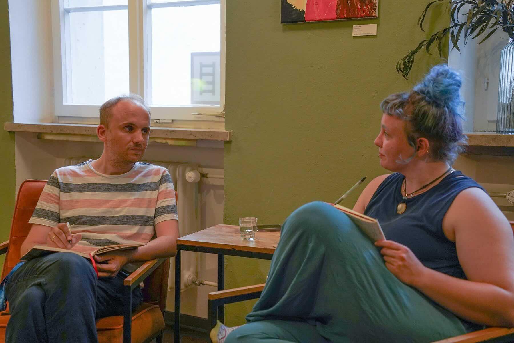 Johannes Frank und eine Besucherin der Schreibgruppe im W1 gemeinsam im Dialog. Dahinter ist eine grüne Wand und eine Pflanze.