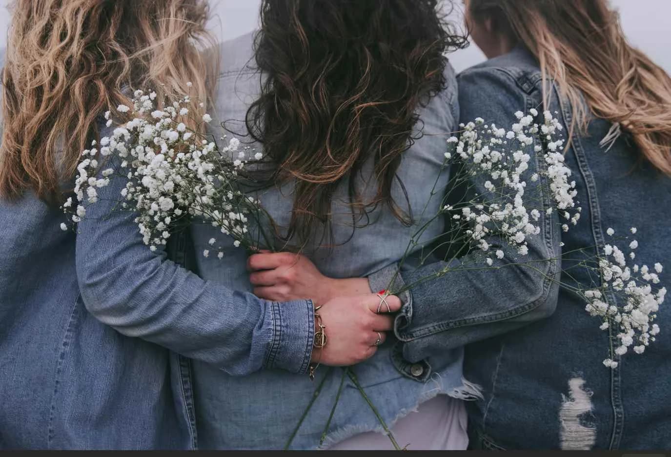 Drei Frauen halten sich in den Armen und sind von hinten mit weißen Blumen zu sehen.