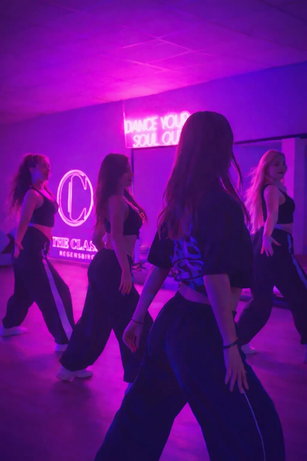Vier junge Frauen tanzen in dem Tanzstudio vor dem Spiegel. An der Wand hängen Neonschilder mit dem Schriftzug "Dance your soul out" und "THE CLASS Regensburg".