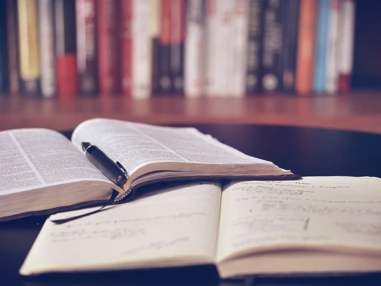Aufgeschlagenes Buch mit Stift und ein Notizheft auf einem Tisch vor einem Bücherregal
