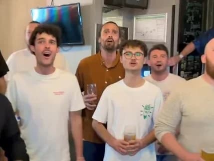 Eine Gruppe Männer singt in einer Kneipe gemeinsam, einige halten Biergläser in der Hand.