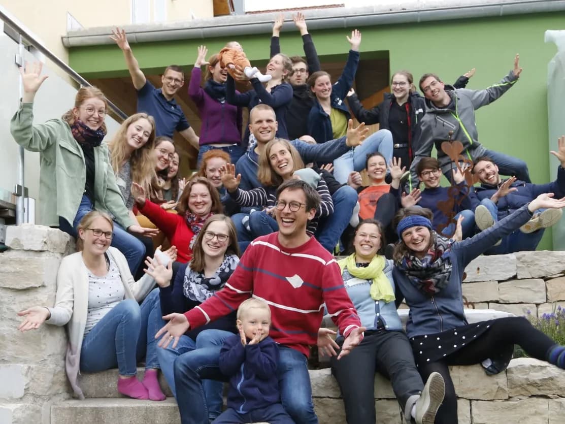Große Chorgruppe, darunter Kinder, sitzt lachend mit erhobenen Armen auf einer Gartentreppe aus Stein.