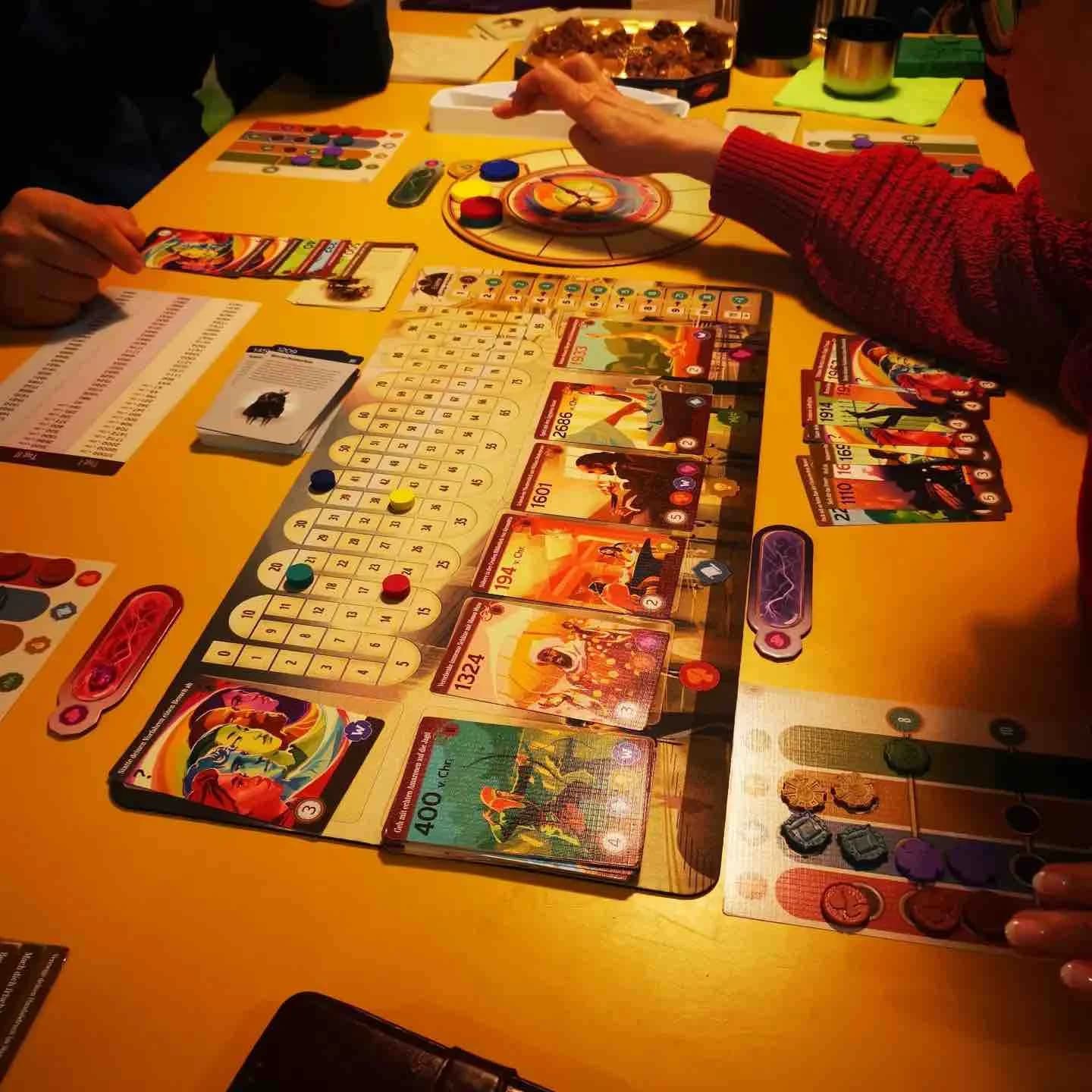 Mehrere Personen spielen an einem Tisch ein Brettspiel mit Karten, Spielsteinen und Punkteleiste.