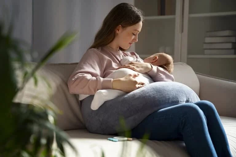 Seitenansicht einer Mutter, die auf der Couch sitzt und ihr Neugeborenes in den Armen hält, repräsentativ für die offene Stillgruppe des Familienzentrums Herztöne.