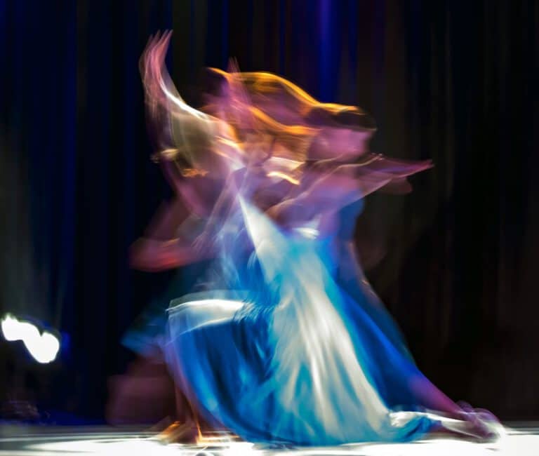 Eine Tänzerin in einem blauen Kleid, die in langsamer Belichtungszeit aufgenommen wurde und deren Bewegungen in einander verschwimmen. Stellvertretend für die "bewegte Meditation".