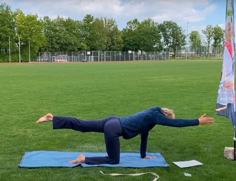 Eine Dame, die im Rahmen von "Sport im Park" eine Pilates-Übung zeigt. Sie streckt auf einer Yoga-Matte ein Bein und eine Hand aus und hält dabei ihren Rücken gerade parallel zum Boden.